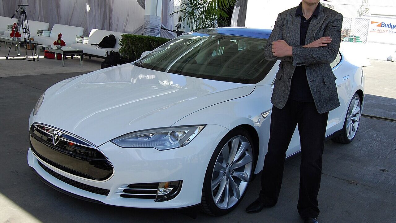 Elon Musk standing beside a white Tesla electric car